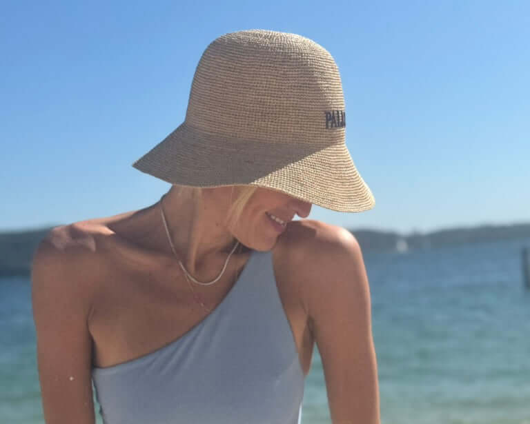 A woman wearing a handwoven sunhat, the PALMA Sunhat, by the water, enjoying a sunny day.