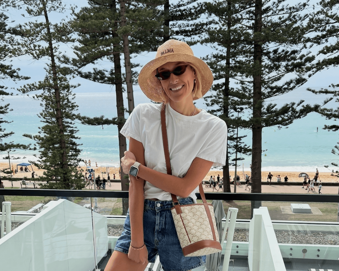 Woman wearing a handwoven sunhat, smiling by the beach with sunglasses and a stylish bag.