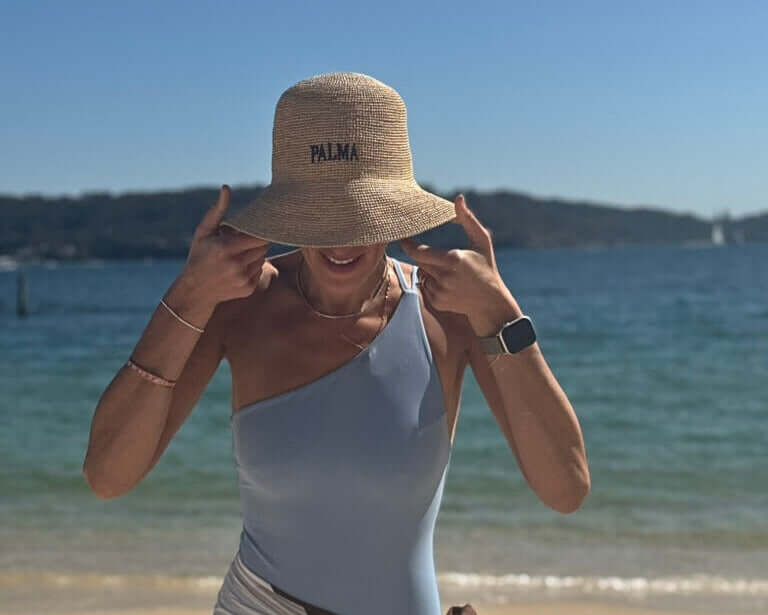A woman gestures playfully while wearing a handwoven sunhat, the PALMA Sunhat, at the beach with water in the background.